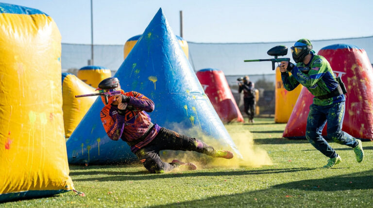 Deux joueurs de paintball en pleine action sur un terrain, un glissant et tirant, l'autre courant et visant. Bunkers colorés et éclaboussures de peinture.