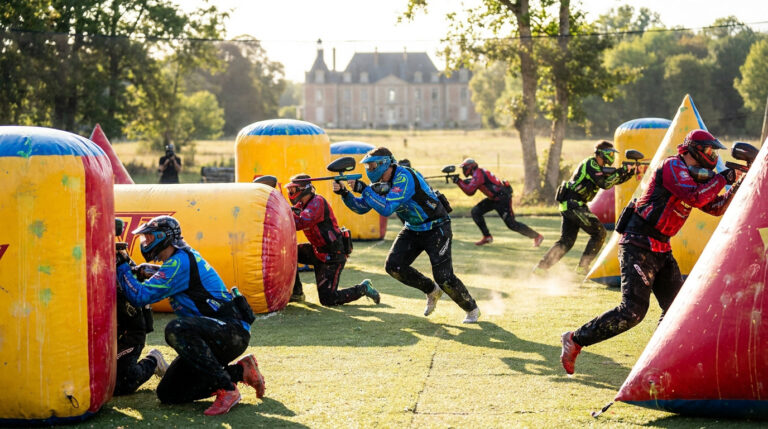 Joueurs de paintball masqués et équipés en pleine action sur un terrain d'obstacles gonflables, avec un château français en fond.